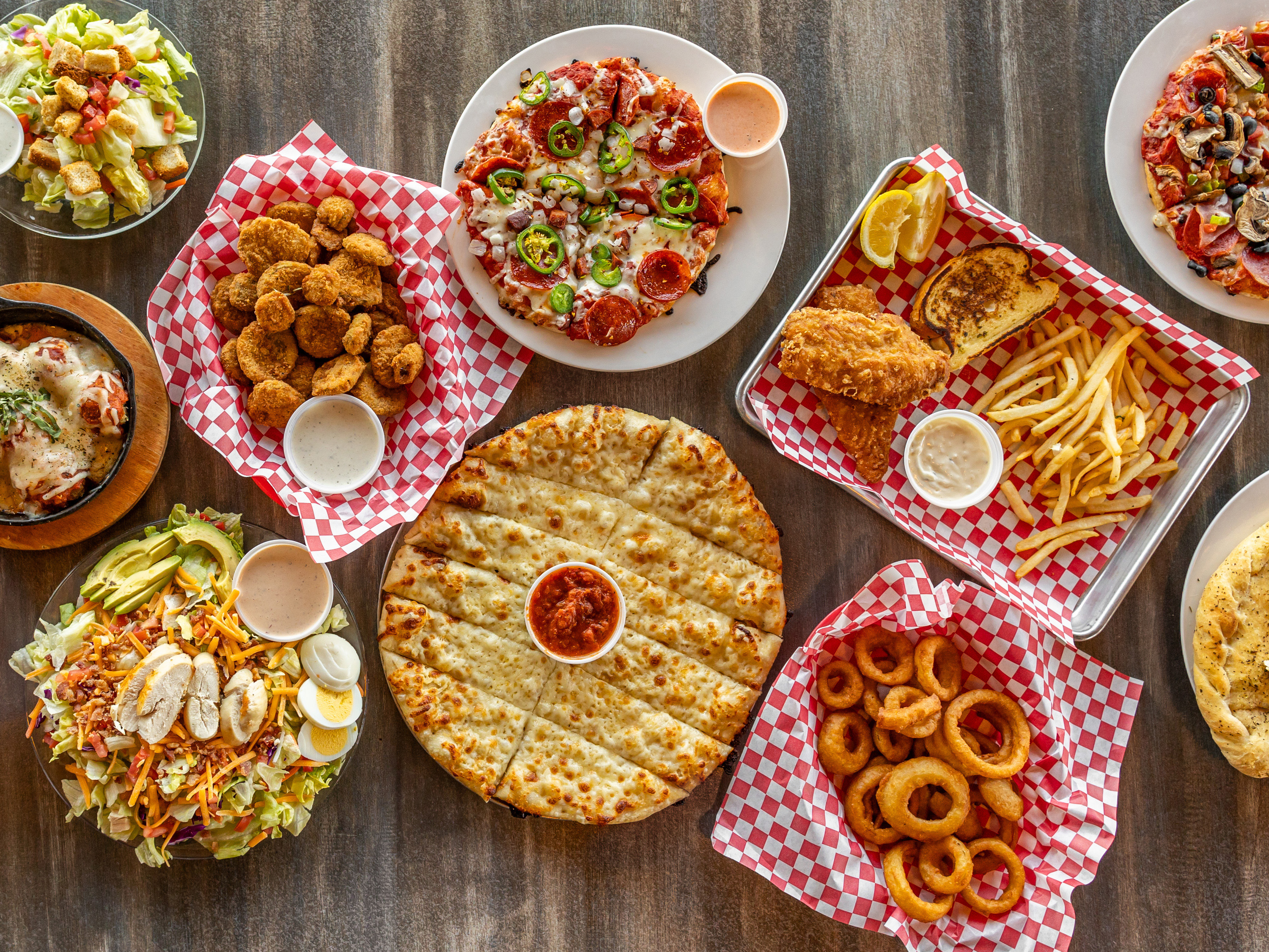 A look down on several dishes of food including salad, breadsticks, onion rings, fried pickles, pizza, and fish & chips