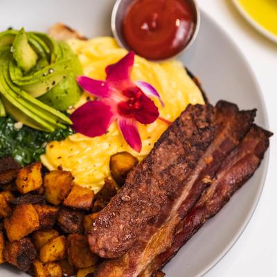 Scrambled eggs with crispy bacon, avocado, breakfast potatoes, and ketchup.