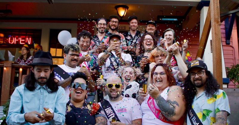 A diverse group of people celebrate joyfully on a porch, wearing colorful outfits and sashes
