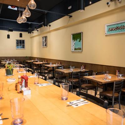 Restaurant interior, with tables set with glasses and utensils, Paintings adorn the beige walls.