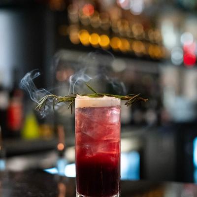 Cocktail in colins glass garnished with burnt rosemary.