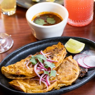 Birria tacos served with lime wedge, sliced onions and radish, alongside a dip.