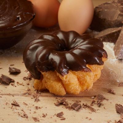 Chocolate cruller pastry on a wooden board with chocolate shavings and eggs in the background.