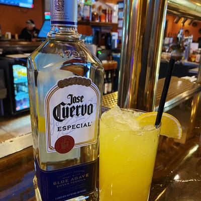 A yellow cocktail garnished with a lemon slice, sits next to a bottle of silver tequila.