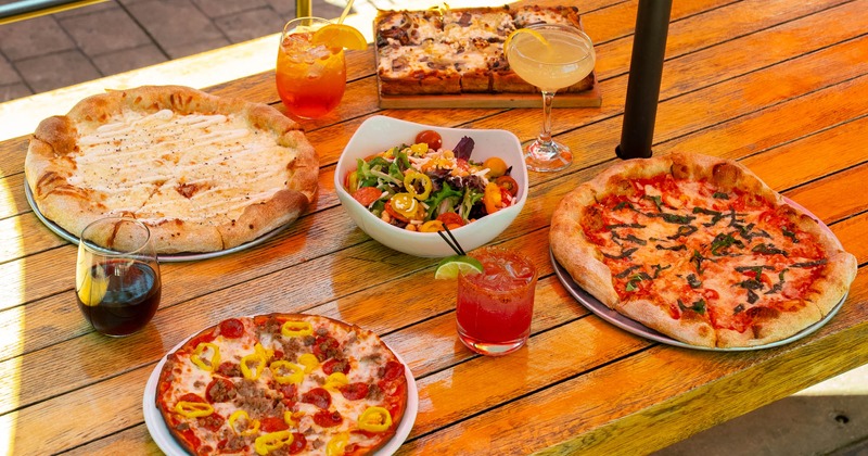 Patio table with assorted pizzas, salad, and cocktails