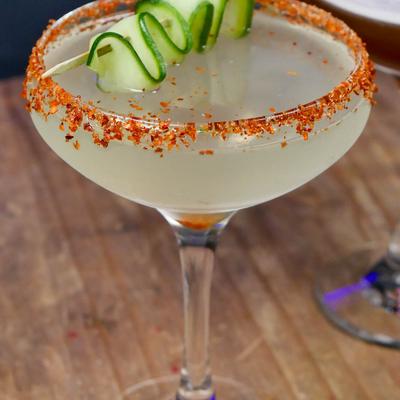 A cocktail in a coupe glass with a chili salt rim and a cucumber curl garnsih.