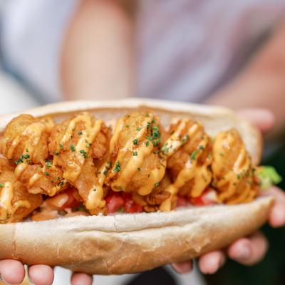 Hands holding a shrimp po' boy sandwich drizzled with red pepper aioli