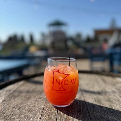 A refreshing orange cocktail in a glass with NC Winery logo, sits on a wooden barrel outdoors.