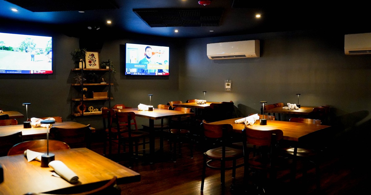 Dimly lit restaurant interior with wooden tables, two wall-mounted TVs, and decorative shelves