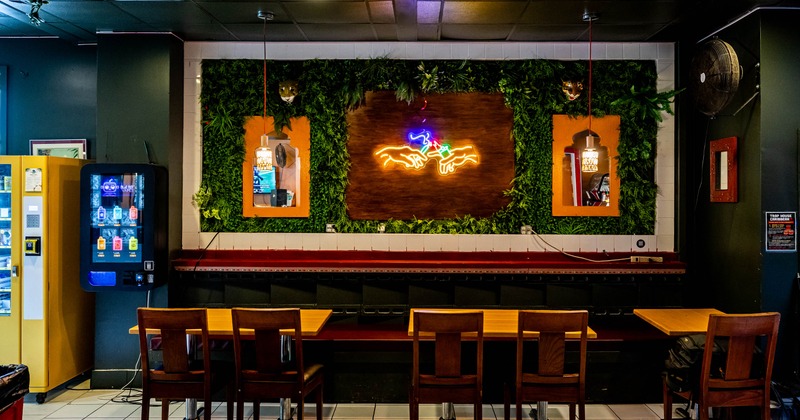 Interior seating area with a foliage covered wall, central neon hand sign, wooden tables and chairs
