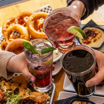 Three friends toasting with colorful drinks over a table filled with onion rings and nachos.