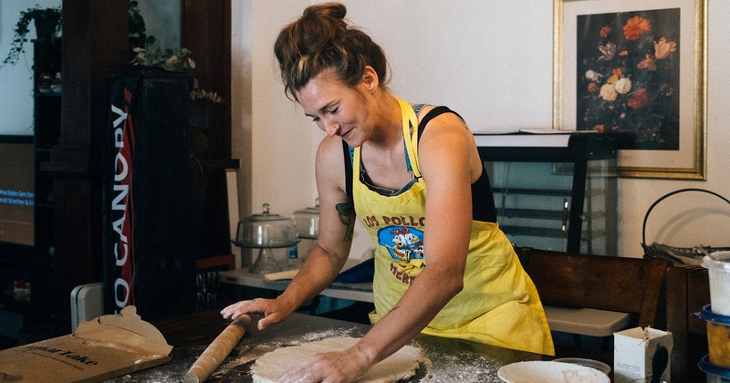 An employee  in a yellow apron smiles while rolling dough on a floured table