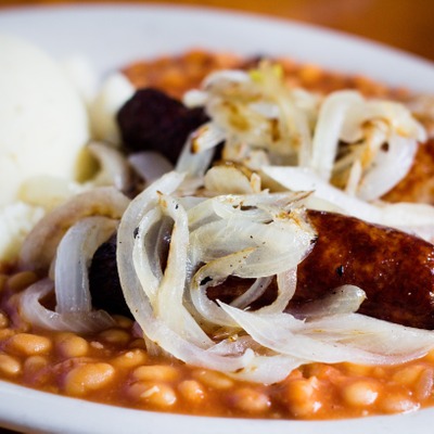 Banger sausages topped with grilled onions on top of baked beans and next to mashed potatoes