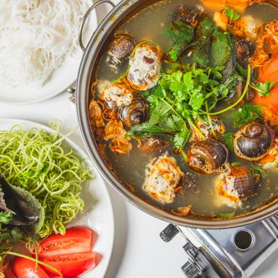 Snail hot pot served alongside a plate of noodles, vegetables and sliced tomatoes.