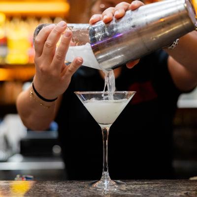 Bartender pouring a martini from a metal shaker into a stemmed glass at the bar.