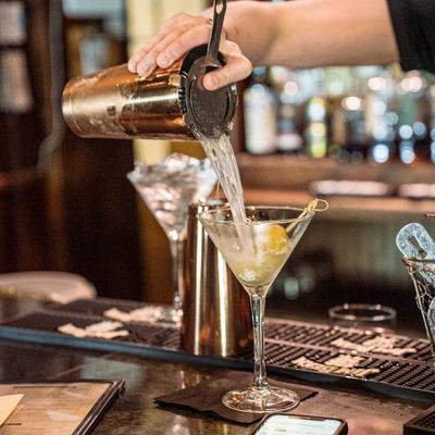 A bartender pouring a cocktail from a shaker into a martini glass with olives.