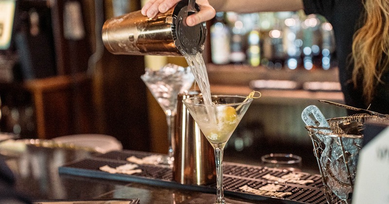 A bartender pouring a cocktail from a shaker into a martini glass with olives