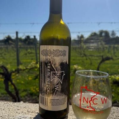 Bottle of NCW Trainwreck white wine, with a wine glass, resting on a stone surface outdoors.