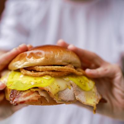 Hands holding a roasted pork sandwich with bacon, fried onions, and pickled green tomato