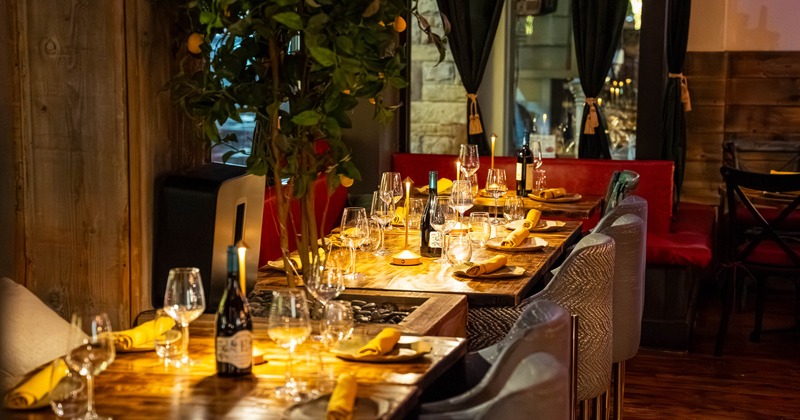 A cozy dining area with wooden tables, wine glasses, candles, yellow napkins and velvet chairs