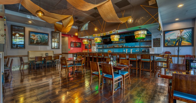 Interior of a restaurant with wooden floors, tables, and chairs, a bar with stools, pendant lighting