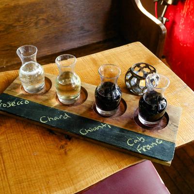 A wooden flight tray with four wine carafes on a table, next to a candle holder