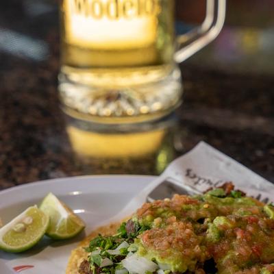 Carne asada taco with guacamole, salsa, cilantro, lime wedges, and a beer.