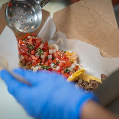 An employee assembles a taco box, adding Pico de Gallo with a ladle.