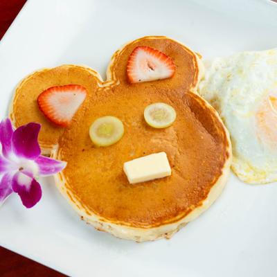 Mickey Mouse-shaped pancake served with a fried egg, butter and fruit garnishes.