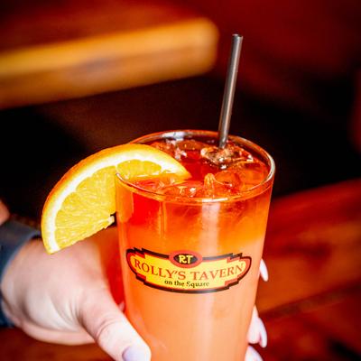 An orange cocktail in a Rolly's Tavern glass with ice and an orange slice garnish.