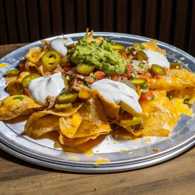 Loaded nachos on a metal tray, topped with sour cream, pulled meat, guacamole, and jalapenos.