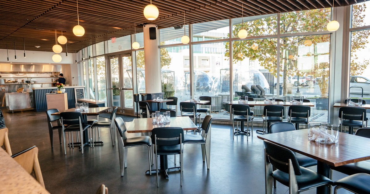 Interior of a modern restaurant with large windows, wooden tables, and dark chairs