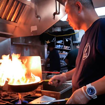 Chef in a busy kitchen, focused as a large flame rises from a pan.