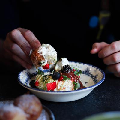 A hand dipping a pastry into a bowl of colorful fruit, cream, and berries.