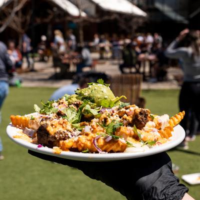 A gloved hand holds a plate of Asada fries, blurred  outdoor setting with people in the background.