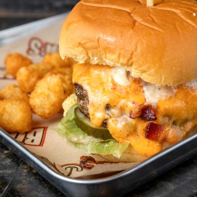Pimento crunch burger topped with pickles and bacon, served alongside tater tots.