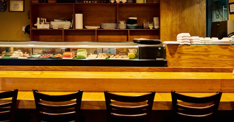 Interior view of a warmly lit sushi bar with a glass food display, wooden shelving