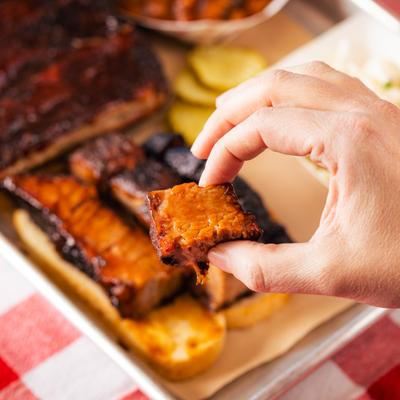 A hand holds a glazed slice of barbecue meat above a tray with ribs.
