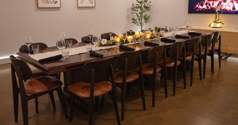 Long dining table set for a meal with glassware, black napkins, white flowers, candles, and chairs in a warmly lit room