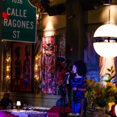Singer holding the microphone during live performance at Calle Dragones, colorful artwork in the background.