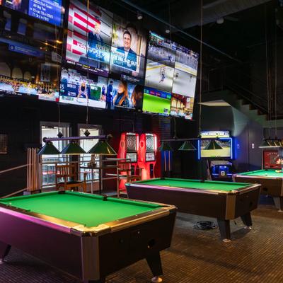 Game room with a row of pool tables and large wall screens.