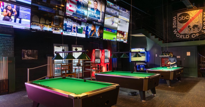 Game room with a row of pool tables and large wall screens
