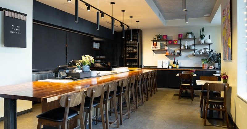 Wine bar- a wooden counter, rows of bar stools, shelves of glassware & bottles, plants, tables