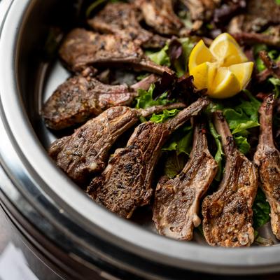 Lamb chops arranged over greens with lemon garnish in a metal bowl.