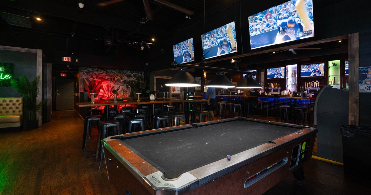 Interior view of a bar featuring a pool table in the foreground, a long bar counter with stools