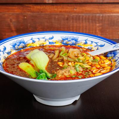Spicy noodle soup with bok choy, peanuts, and chili oil in a blue floral bowl.
