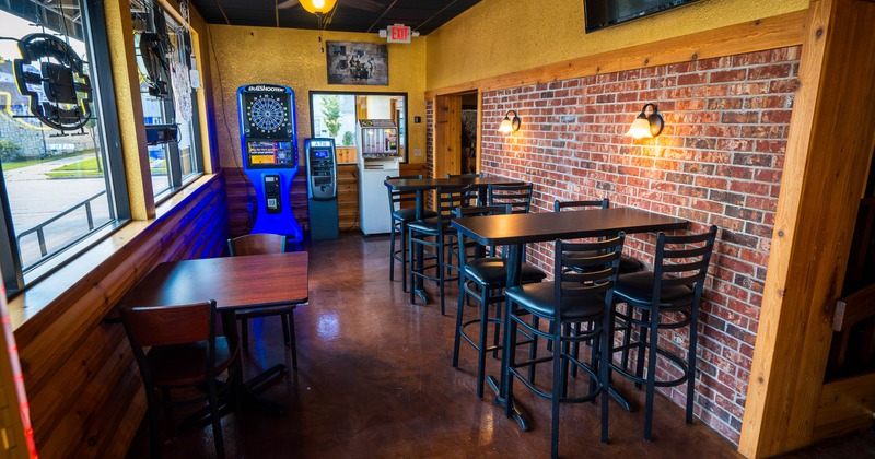 Interior of a warmly lit bar or restaurant with tables, chairs, a brick accent wall, windows, an electronic dartboard