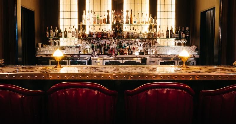 A dimly lit bar with a marble countertop, red leather bar stools, and liquor bottles