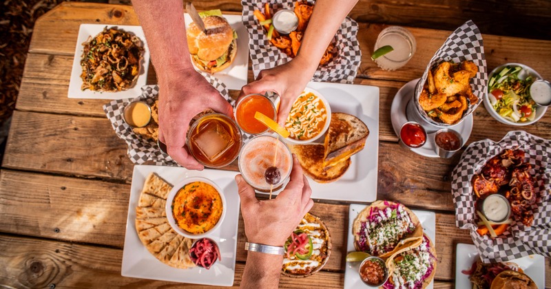 Overhead view, three people toast with drinks at a picnic table filled with assorted food dishes