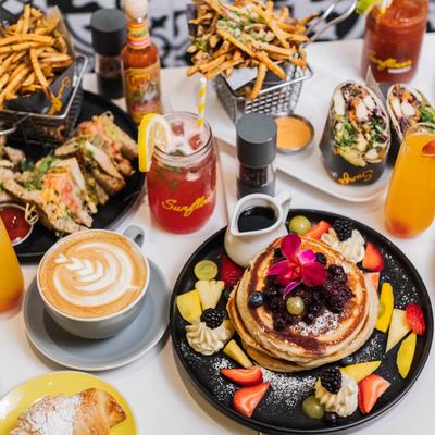 Colorful brunch spread with pancakes, fruit, sandwiches, coffee, and cocktails.